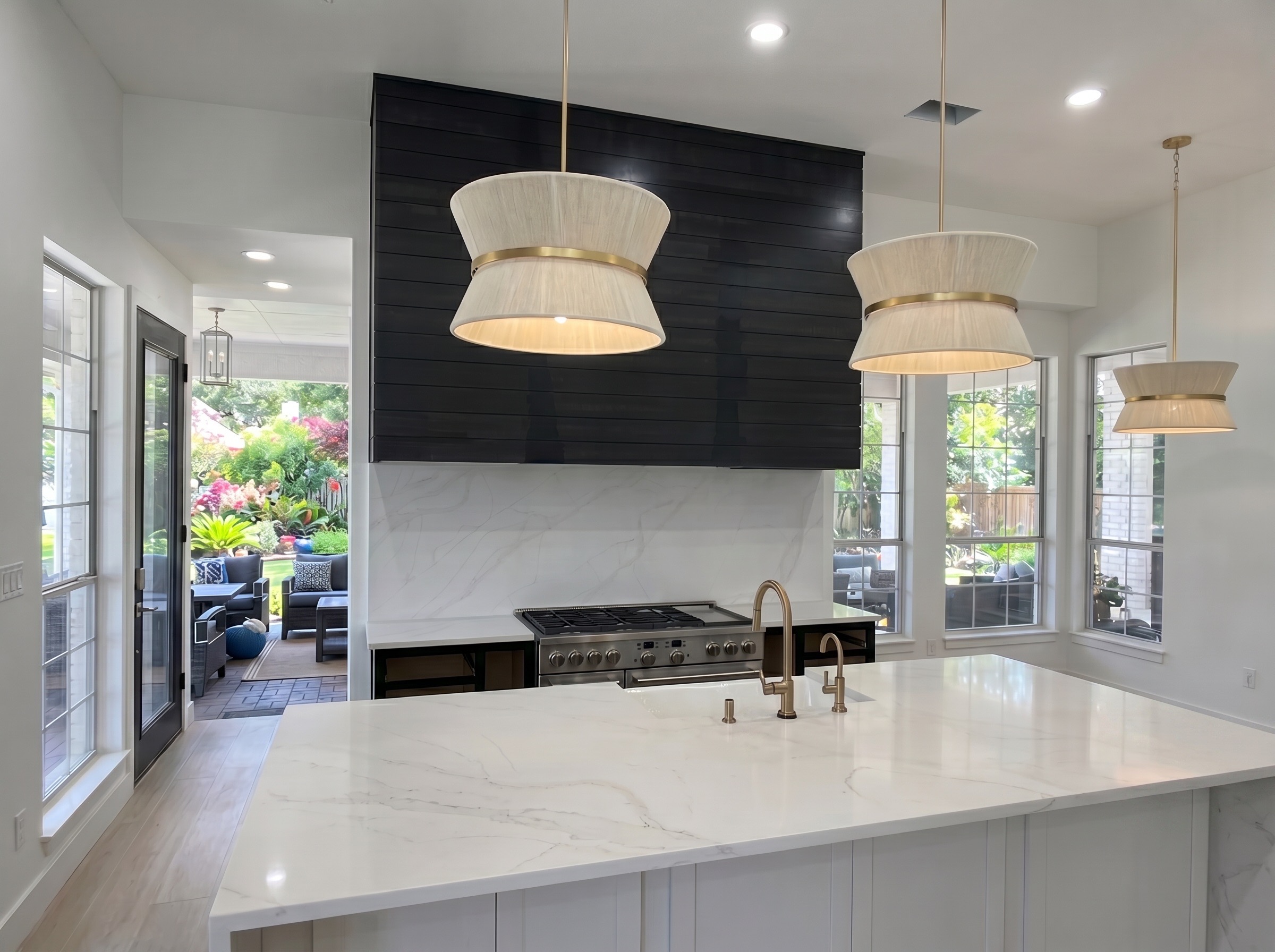 Bright remodeled kitchen with a white marble waterfall island, black shiplap range hood, brass pendant lighting, and a view to the garden