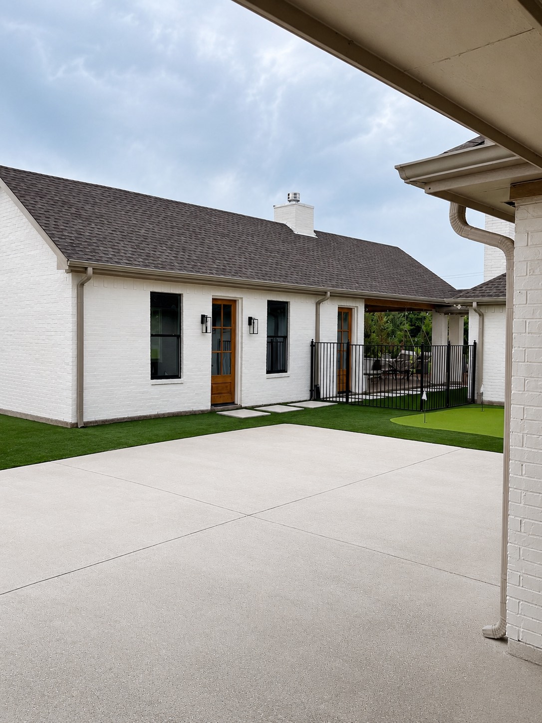 White-brick home exterior with a covered patio, broom-finish concrete, artificial turf lawn, and a backyard putting green