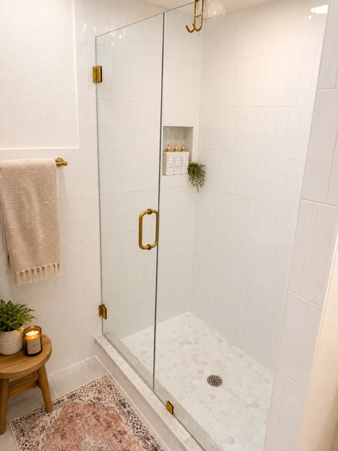 Walk-in shower with white vertical tile, frameless glass, brushed brass fixtures, a hex-tile floor, and a niche styled with greenery